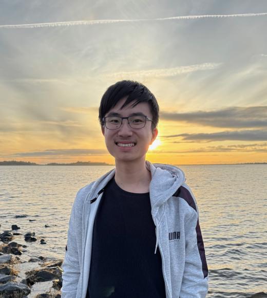 Smiling person with glasses by rocky shoreline at sunset, wearing a light jacket
